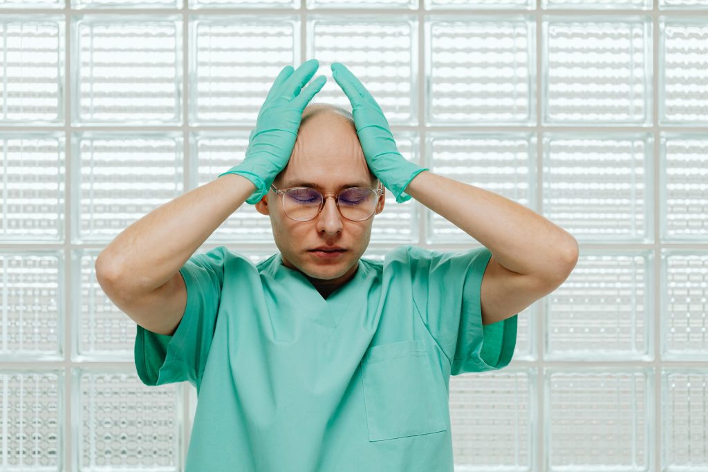 A healthcare worker in green scrubs looking stressed, hands on head with eyeglasses.